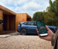 Das Foto zeigt eine Hand mit einem Smartphone im Vordergrund, das Display zeigt die Solarwatt-App. Im Hintergrund unscharf rechts ein Privathaus als Flachbau mit umlaufender Holzverkleidung, ein blaues Auto an eine Wallbox angeschlossen.