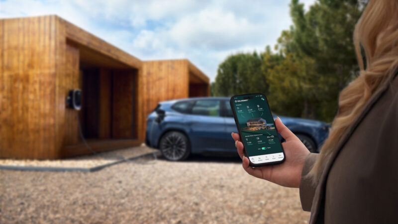 Das Foto zeigt eine Hand mit einem Smartphone im Vordergrund, das Display zeigt die Solarwatt-App. Im Hintergrund unscharf rechts ein Privathaus als Flachbau mit umlaufender Holzverkleidung, ein blaues Auto an eine Wallbox angeschlossen.