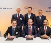Gruppe von Männern in schwarzen Anzügen hinter einem Tisch vor Firmenlogos - Vertragsunterzeichnung für Gleichstrom-Konverter.