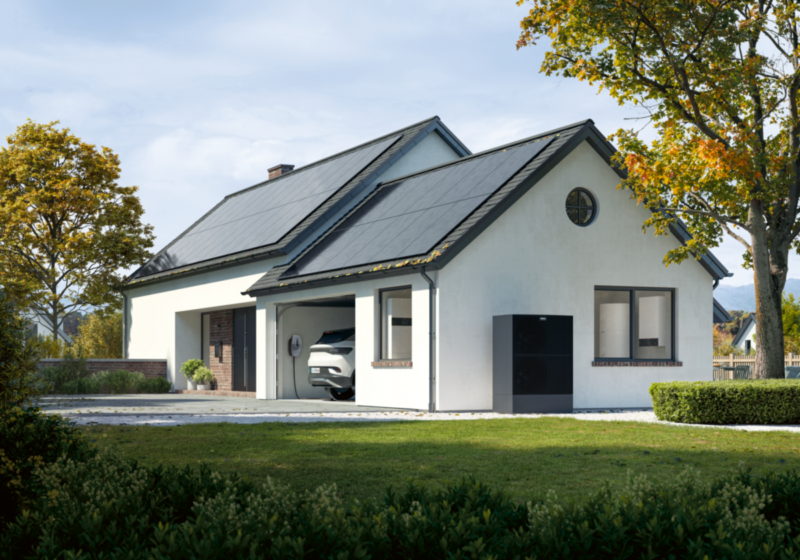 Das Bild zeigt ein weißes Einfamilienhaus, schräg von der Seite, rechts und links Bäume, mit Blick aus dem Grün, in der Garage ein PKW mit Wallbox und vor der Garage eine Wärmepumpe.