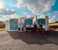 16 Personen vor einem Batteriespeicher, der in Containern untergebracht ist, unter blauem Himmel mit Wolken.