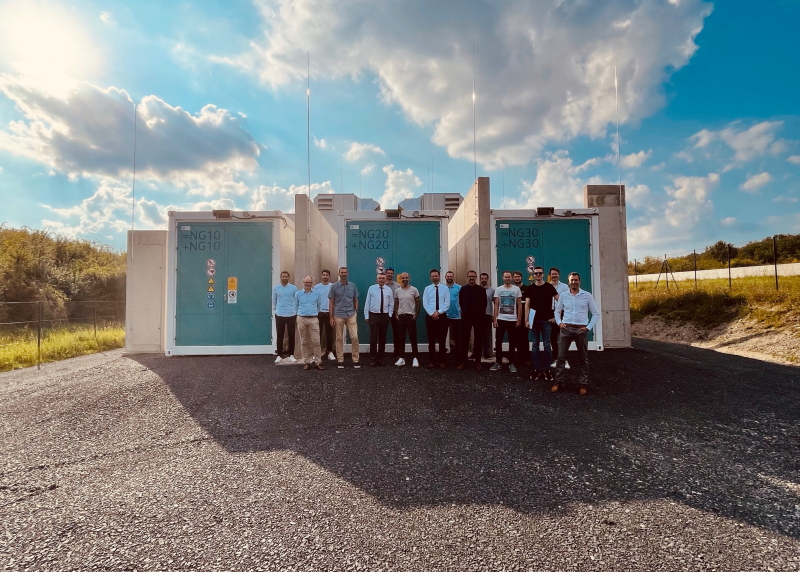 16 Personen vor einem Batteriespeicher, der in Containern untergebracht ist, unter blauem Himmel mit Wolken.