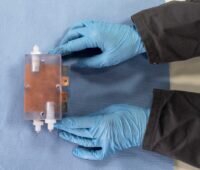 Zu sehen ist eine Batteriezelle der Iron-Redox-Flow-Stromspeichertechnologie, gehalten von Händen in blauen Handschuhen.