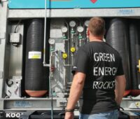 Ein Mann im Rücken zur Kamera, auf seinem T-Shirt ist "Green Energy Rocks" zu lesen, vor einer Wasserstoffeinheit.