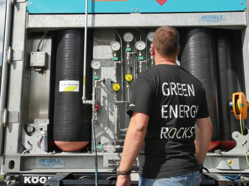 Ein Mann im Rücken zur Kamera, auf seinem T-Shirt ist "Green Energy Rocks" zu lesen, vor einer Wasserstoffeinheit.