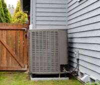 Luft-Wärmepumpe mit wenig Abstand: Das Bild zeigt am linken Rand einen Gartenzaun, dicht daneben eine Wärmepumpe an der Hauswand.