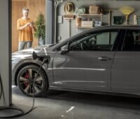 Ein Auto in einer Garage lädt an einer Wallbox, ein Mann im Hintergrund tippt auf sein Smartphone - Laden mit Solar-Strom.