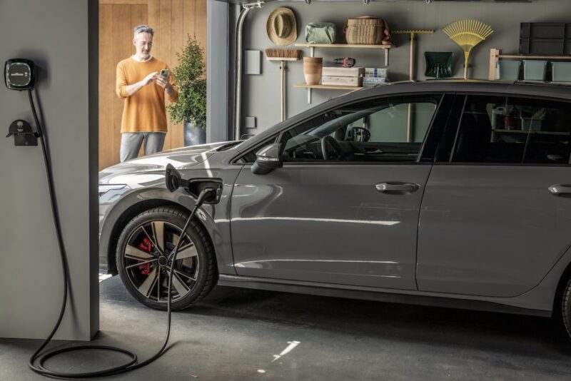 Ein Auto in einer Garage lädt an einer Wallbox, ein Mann im Hintergrund tippt auf sein Smartphone - Laden mit Solar-Strom.