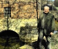 Bernd Wallters, rechts im Bild sammelt Wasserkraftwerke. Er steht vor einem historischen Backsteingebäude, einer Wasserkraftanlage über einem Kanal.