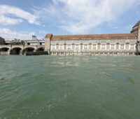 Ein altes Wasserkraftwerk am Rhein und bviel Wasserim Vordergrund.