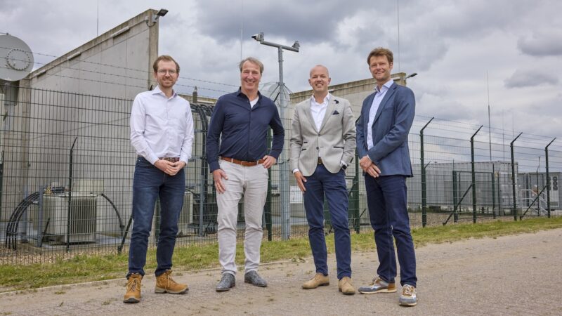 Vier Männer stehen vor einer eingezäunten Hochspannungsanlage mit Transformatoren und weiteren elektrischen Anlagen.