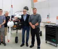 Vier Mitglieder des THWS-Projektteams stehen in einer Werkhalle vor dem zweisitzigen Reisemotorsegler „Taifun 17 H₂“. Die Verkleidung des Flugzeugmotors ist geöffnet und gibt den Wasserstoff-Elektroantrieb frei. Rechts im Bild stehen ein Werkstattwagen und ein Roll-up zur mobilen Wasserstoff-Tankstelle.
