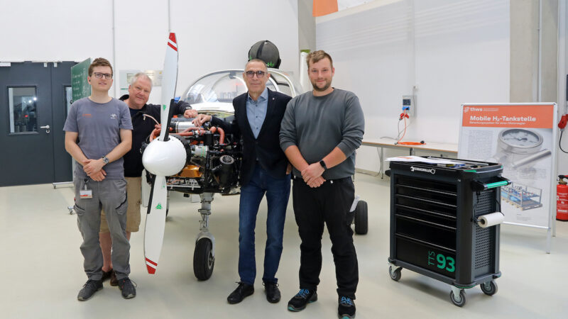 Vier Mitglieder des THWS-Projektteams stehen in einer Werkhalle vor dem zweisitzigen Reisemotorsegler „Taifun 17 H₂“. Die Verkleidung des Flugzeugmotors ist geöffnet und gibt den Wasserstoff-Elektroantrieb frei. Rechts im Bild stehen ein Werkstattwagen und ein Roll-up zur mobilen Wasserstoff-Tankstelle.