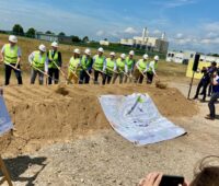 Männer mit gelben Warnwesten und Helmen und Schaufeln stehen hinter Erdwall, auf dem ein Banner liegt, Fotografen - Baustart für das Wasserstoff-Startnetz