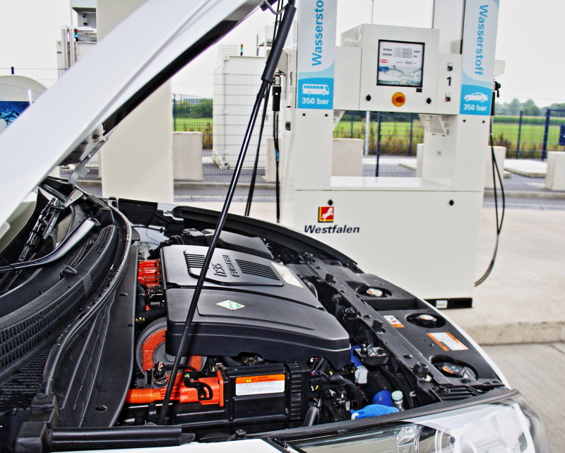 Blick auf die geöffnete Motorhaube eines Brennstoffzellen-PkWs vor einer Wasserstoff-Zapfsäule.