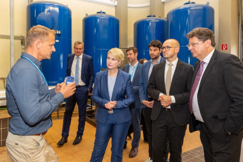 Sechs Männer und eine Frau vor blauen Behältern - Geothermie-Anlage in Neustadt-Glewe.