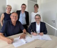 Fünf Personen stehen und sitzen an einem Besprechungstisch in einem Büro. Zwei Männer unterzeichnen Vertragsunterlagen, während drei weitere Personen, zwei Frauen in der Mitte ein Mann, hinter ihnen stehen.