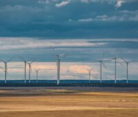 Eine Reihe großer Windenergie-Anlagen vor Gewitterhimmel, im Vordergrund ein Stoppelfeld. Symbolbild für Onshore-Windenergie, Windpark.