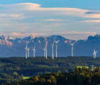 Das Foto zeigt aus der Luft von vorne sonnenbeschienene Windräder im Wald vor diesigem Alpenpanorama mit aufziehender Bewölkung darüber.