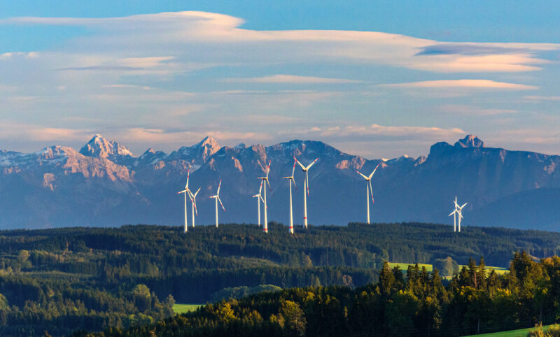 Das Foto zeigt aus der Luft von vorne sonnenbeschienene Windräder im Wald vor diesigem Alpenpanorama mit aufziehender Bewölkung darüber.