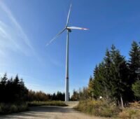 Windenergie-Anlage vor blauem Himmel, davor ein Weg und Wald aus Nadelbäumen.