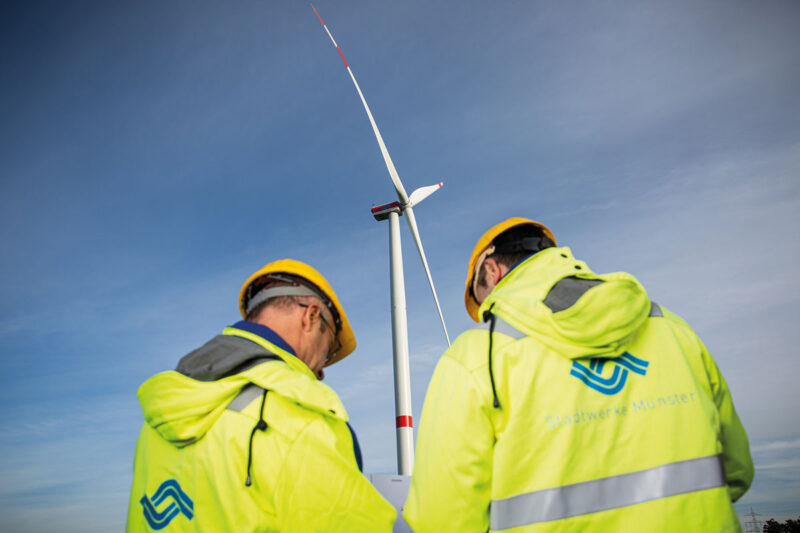 Männer in gelber Arbeitsschutzkleidung vor einer Windenergie-Anlage.