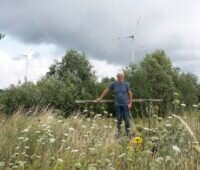 Johannes Maibom steht auf einer Wiese, die die Gesellschaft Reuthwind für die Photovoltaik nutzen möchte. Im Hintergrund ist der Windpark zu sehen. Maibom hält die bayerische 3D-Regel nicht für sinnvoll.