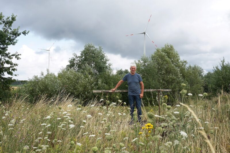 Johannes Maibom steht auf einer Wiese, die die Gesellschaft Reuthwind für die Photovoltaik nutzen möchte. Im Hintergrund ist der Windpark zu sehen. Maibom hält die bayerische 3D-Regel nicht für sinnvoll.