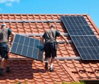 Im Bild zwei Männer, die ein Photovoltaik-Modul auf ein Dach schleppen, die Initiative SolAixQ ⎼ Solar lernen im Aachener Quartier bildet Solarhelfer:innen aus.