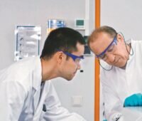 Zwei Männer mit Schutzbrillen arbeiten an einem Glaskolben, in dem eine grüne Flüssigkeit ist - Material für die Natrium-Ionen-Batterie