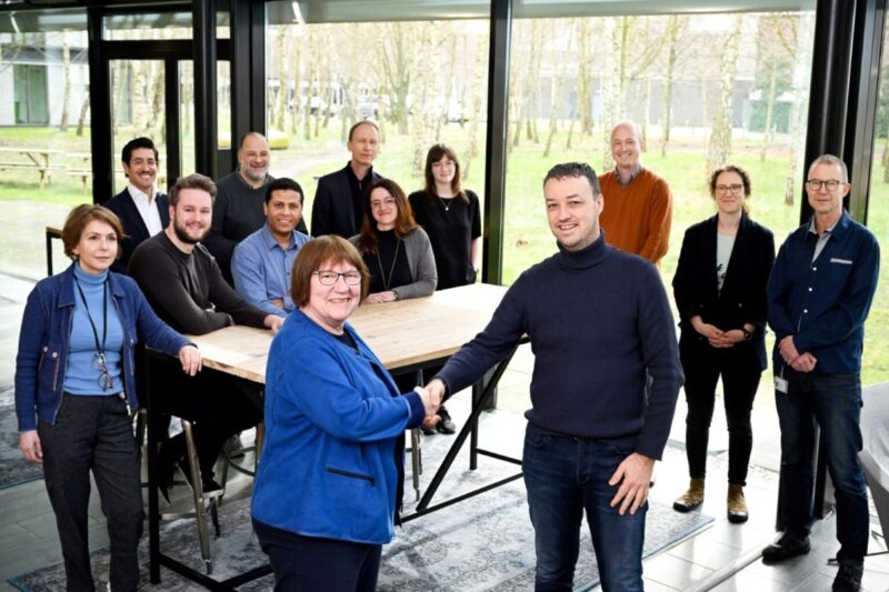 Zwei Personen reichen sich die Hand udn blicken in die Kamera, während drumherum weitere Personen stehen.