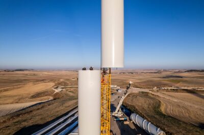 Auf einer Baustelle fügen Kräne zwei Teile eines Windturms ineinander.