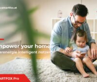 Smartfox: Enpower your home. Solarenergie intelligenz nutzen. Zu sehen ist ein auf einem Teppich sitzender Mann mit Mädchen auf dem Schoß, der auf ein Smartphone blickt und lacht.