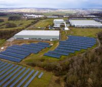 Luftbild einer Fabrik im Vordergrund mit Solaranlagen, im HIntergrund mit Windenergie.