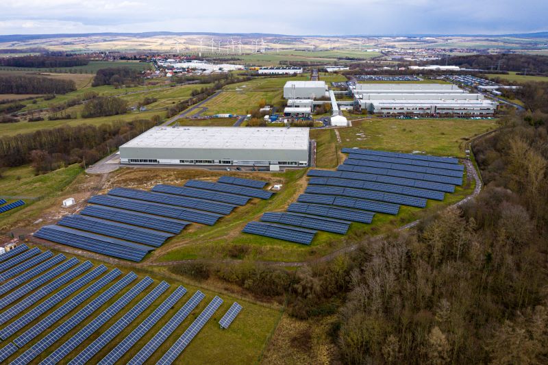 Luftbild einer Fabrik im Vordergrund mit Solaranlagen, im HIntergrund mit Windenergie.