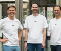 Drei Männer in Jeans und weißen T-Shirts stehen in den Geschäftsräumen nebeneinander und blicken lächelnd in die Kamera.