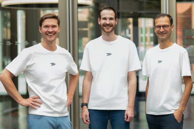 Drei Männer in Jeans und weißen T-Shirts stehen in den Geschäftsräumen nebeneinander und blicken lächelnd in die Kamera.