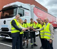 Mehrere Personen in neongelben Warnwesten stehen vor einem batterieelektrischen Mercedes-Benz eActros 600 mit hylane- und Coca-Cola-Schriftzug an der Kabine und stoßen vor dem Fahrzeug mit Getränkeflaschen an.