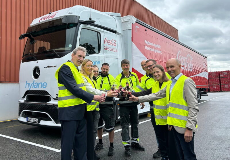 Mehrere Personen in neongelben Warnwesten stehen vor einem batterieelektrischen Mercedes-Benz eActros 600 mit hylane- und Coca-Cola-Schriftzug an der Kabine und stoßen vor dem Fahrzeug mit Getränkeflaschen an.