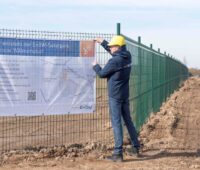 Ein Bauarbeiter mit Helm installiert ein Infoschild an einem Bauzaun. Das informiert über den geplanten Solarpark.