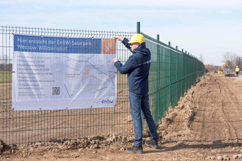 Ein Bauarbeiter mit Helm installiert ein Infoschild an einem Bauzaun. Das informiert über den geplanten Solarpark.