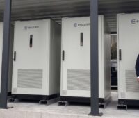 Zwei Personen mit Schutzhelmen neben vier großen Batteriespeichern.