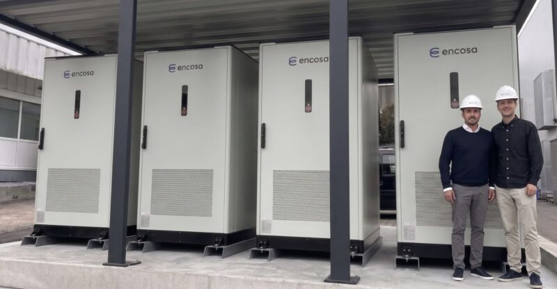 Zwei Personen mit Schutzhelmen neben vier großen Batteriespeichern.