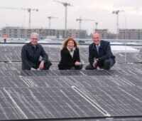 Eine Dame, zwei Herren in Business-Outfit knien lächelnd auf einem Dach zwischen Solarmodulen.