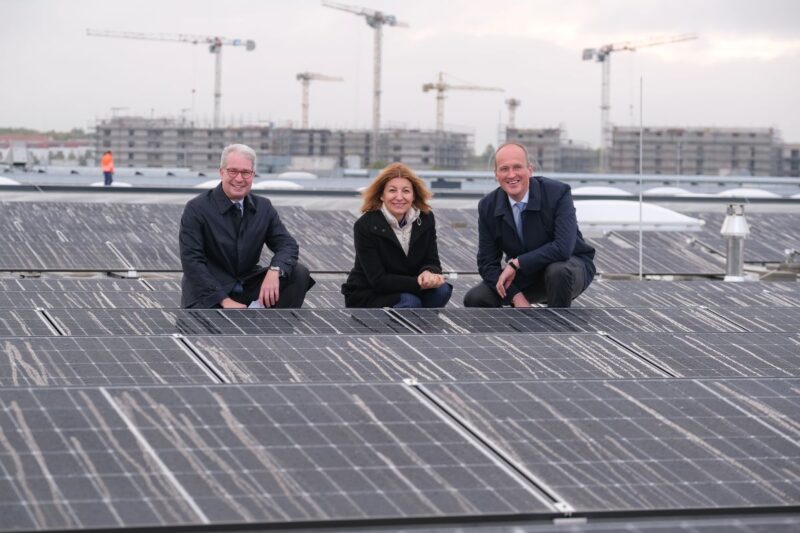 Eine Dame, zwei Herren in Business-Outfit knien lächelnd auf einem Dach zwischen Solarmodulen.