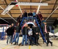 Gruppe von Männern und Frauen auf Leiter in einer Halle, darüber die Flugwindkraft-Anlage von Enerkite.