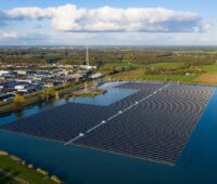 Solarmodule schwimmen auf einem See in den Niederlanden.. Links vom See befindet sich ein Industriegebiet, im Hintergrund Grünland.