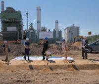 6 Personen vollziehen einen Spatenstich vor Anlagen der Chemieindustrie.