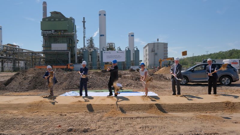 6 Personen vollziehen einen Spatenstich vor Anlagen der Chemieindustrie.