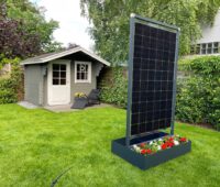 Ein hochkant stehendes Solarmodul mit Blumenkasten vor einem Gartenhäuschen.